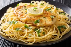 a black plate topped with pasta and chicken covered in parmesan cheese, garnished with lemon wedges