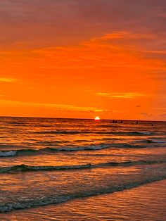 A view of the sunset at the shore from the beach. The sky is a bright orange with half of the sun still shining. Soft waves are flowing onto the shore. The waves have a bit of the orange reflected onto them from the sunset. Golden Hour At The Beach, Orange Ocean Aesthetic, Orange Sunset Beach, Sunrise Orange, Ocean Surf, Orange Sunset, Look At The Sky