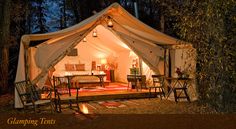 a tent set up in the woods at night with lights on and chairs around it