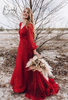 a woman in a long red dress holding a bouquet and standing next to a tree