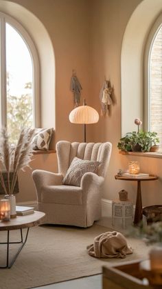 A cozy living room corner. A plush, beige armchair with a textured fabric sits near a round wooden table adorned with candles and a book. Above the chair, two decorative items hang on the wall, and a floor lamp with a unique design illuminates the space. To the left, a window offers a view of trees outside, and a potted plant sits on the windowsill. The room's walls are painted in a soft beige hue, and a rug with a muted tone covers the floor. Cozy Living Room Corner, Round Wooden Table, Beige Armchair, Neutral Paint Color, Living Room Corner, Christmas Decorations Living Room, Christmas Living Rooms