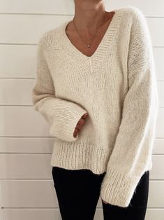 a woman standing in front of a white brick wall wearing a sweater and black pants