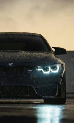 the front end of a black bmw car with headlights on in the rain at night