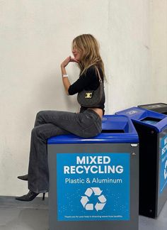 a woman sitting on top of a blue recycling bin