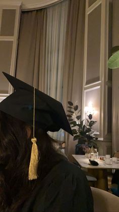 a woman wearing a black graduation cap and gown in a room with white walls, plants and curtains