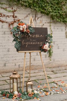 a welcome sign with flowers and greenery on the ground next to a brick wall