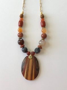 Lightweight oval wooden pendant has different shades of brown. Necklace has different shapes and sizes of stone beads in earthtone colors and hangs from a gold plated chain with oblong links and has a gold plated lobster clasp. Brown Wooden Beads Oval Jewelry, Brown Wooden Oval Beads Jewelry, Brown Jewelry With Oval Wooden Beads, Bohemian Brown Necklace With Oval Beads, Brown Wooden Beads Pendant Necklace, Brown Natural Stone Oval Bead Jewelry, Handmade Brown Oval Necklace, Brown Oval Bead Jewelry With Natural Stones, Brown Jewelry With Oval Natural Stones