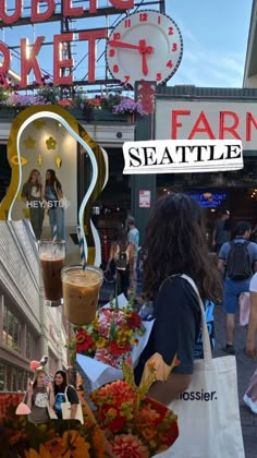 people are shopping at an outdoor market with flowers and drinks in front of the sign