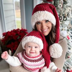 a woman holding a baby wearing a red and white knitted hat with pom poms