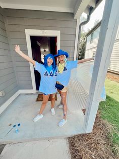 two people in blue shirts and hats standing on the front porch of a house with their arms outstretched