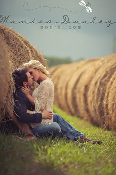 a man and woman sitting in front of hay bales on the grass with their arms around each other