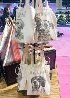 totes hanging on display at an event with people in the background and pink carpet