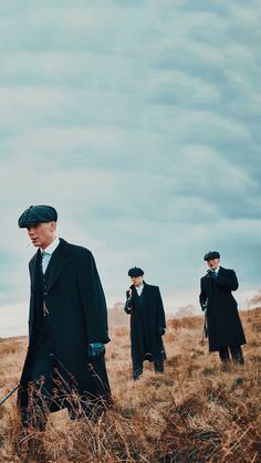 three men in black coats and hats are walking through the grass with one man holding a cane