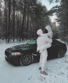 two people standing next to a black car in the snow with trees behind them and one person wearing white