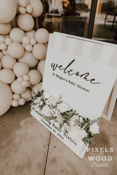 a welcome sign with white flowers and greenery on the sidewalk in front of balloons