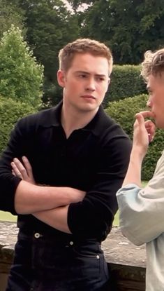 two young men standing next to each other in front of some bushes and trees with their arms crossed