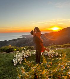 a man and woman kissing in front of the sunset on top of a grassy hill