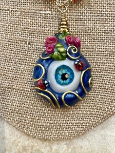 "Let the haters know you know their intentions with this over the top, more is more, evil eye nazar pendant. Even the two ladybugs standing guard beside the two tiny bouquets of fuscia roses are keeping watch over you. Large, dark blue pearlescent evil eye pendant is embellished with gold colored wire flourishes.  Every polymer clay evil eye pendant is sculpted and painted from scratch, starting with handcrafted, realistic-ish eye cabochons that have been marbled with dark blue, light blue, and Unique Evil Eye Jewelry Gift, Unique Evil Eye Jewelry As A Gift, Eye-catching Handmade Jewelry For Gifts, Unique Evil Eye Jewelry For Festival, Adjustable Teardrop Evil Eye Jewelry, Unique Evil Eye Necklace For Gift, Unique Eye-shaped Jewelry For Gifts, Eye-catching Evil Eye Jewelry For Gift, Healing Evil Eye Amulet Jewelry