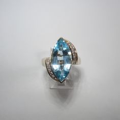 "A large marquise cut Blue Topaz sits in the center of this 14k yellow gold setting. The ring is adorned with natural diamond accents on either side. Ring Size 7.5. Blue Topaz - 5.5 Carat (18mm x 9mm x 5.9mm) Diamonds - There are 16 total diamonds, about 1.5mm round each. Markings - \"14K\" and a maker's mark (pictured). Weight - 6.3 grams This an estate piece in excellent condition. It has been professionally cleaned and sanitized for your peace of mind. The Trading Post is based in Chardon, Oh Blue Marquise Topaz Ring For Anniversary, Marquise Cut Topaz Ring For Anniversary, Marquise Blue Topaz Ring With Accent Stones, Formal Marquise-cut Blue Topaz Ring, Blue Marquise Topaz Ring, Formal Marquise Topaz Ring With Accent Stones, Marquise Blue Topaz Ring, Blue Diamond Marquise Ring With Accents, Marquise Diamond And Topaz Ring