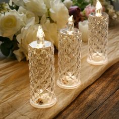 three glass candles sitting on top of a wooden table next to white flowers and greenery