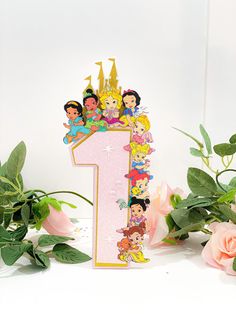 there is a pink number one with princesses on it next to flowers and greenery