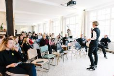 a woman standing in front of a room full of people sitting down and talking to each other