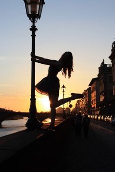 a woman is dancing on the street in front of a lamp post with a sunset behind her