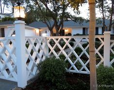 a white fence in front of a house with trees and bushes around it at dusk