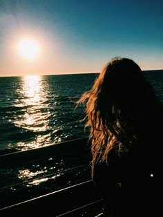 a woman is looking out at the ocean as the sun sets over the water with her hair blowing in the wind