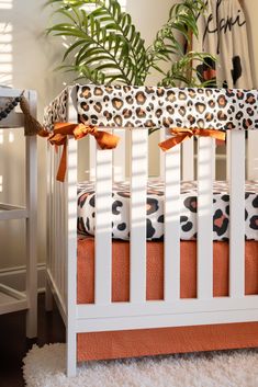 a white crib with orange and black bedding in a room next to a potted plant