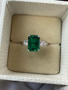 an emerald and diamond ring sits in a white box on top of a cloth covered surface