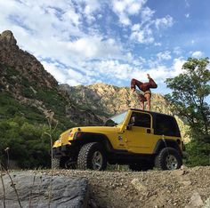 a man standing on top of a yellow jeep in front of some rocks and mountains