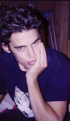 a young man sitting on top of a bed with his hand under his chin and looking at the camera