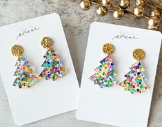 two pairs of colorful christmas tree earrings sitting on top of a white card next to gold balls