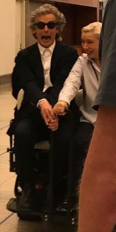 an older man in a suit and sunglasses sitting on a wheel chair with another person standing next to him