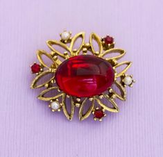 Above is a gold tone brooch with faux ruby gem at the center. --Details-- Brand: NA Color: Gold, Red Length: Next to a ruler in the last image Red Formal Costume Jewelry Brooches, Art Deco Ruby Jewelry As A Gift, Formal Ruby Brooch Jewelry, Red Vintage Lapel Pin For Gift, Vintage Red Lapel Pin As Gift, Vintage Red Lapel Pin For Gift, Luxury Red Art Deco Jewelry, Red Vintage Lapel Pin As Gift, Red Oval Art Deco Jewelry