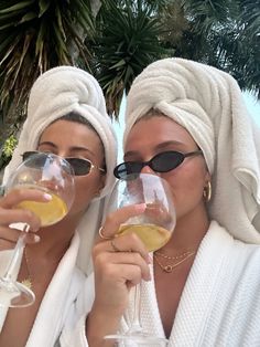 two women in white robes holding wine glasses and looking at the camera with palm trees behind them