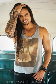 a man with long hair standing in front of a green car wearing a tank top