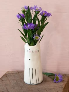 a white vase with purple flowers in it and a cat face painted on the side