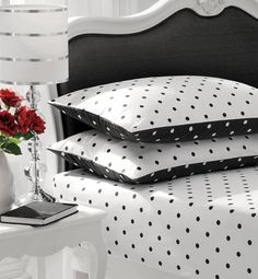 three pillows on a white bed with black and white polka dot print, one has red roses in a vase next to it