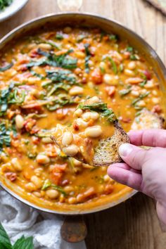 a hand holding a piece of bread over a bowl of beans and spinach soup