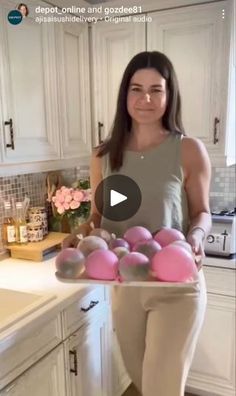 a woman standing in a kitchen holding a tray filled with pink eggs