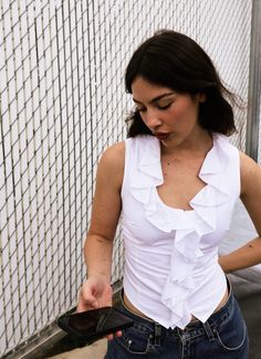 a woman standing next to a fence looking at her cell phone while wearing a white top with ruffles on it