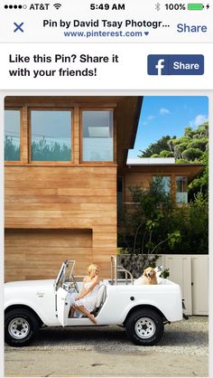 a woman sitting in the back of a white car next to a wooden house and dog