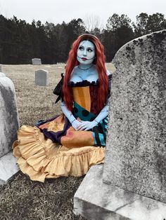 a woman with red hair and makeup sitting on the ground next to a gravestone