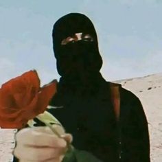 a person with a black mask holding a red rose in their hand and looking at the camera