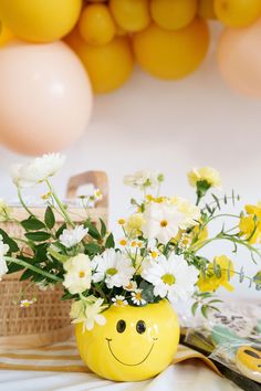 a yellow smiley face vase with daisies and flowers in it on a table next to balloons