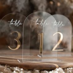 three clear acrylic table numbers sitting on top of a wooden board with gold lettering