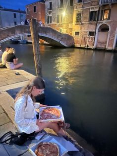 a woman sitting on the edge of a canal with two pizzas in front of her