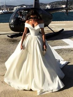 a woman standing in front of a helicopter wearing a white dress with off the shoulder sleeves